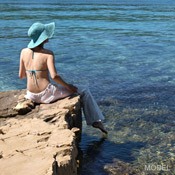 The back of a woman wearing loose white pants, a bikini top, and a blue hat sitting on a rock, dipping her toe in the ocean.