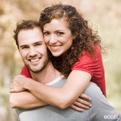 Young woman in red shirt wraps her arms around a young man from behind, both grinning at the camera.