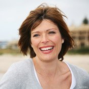 Close-up of woman with short, windblown mahogany hair stands on a beach and laughs facing the camera.