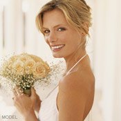 Blonde woman in white halter wedding dress smiles at camera over her bare left shoulder as she holds a bouquet.