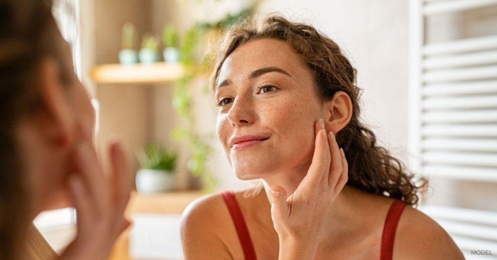 Woman with clear and radiant skin (model) examining her face closely in the mirror.