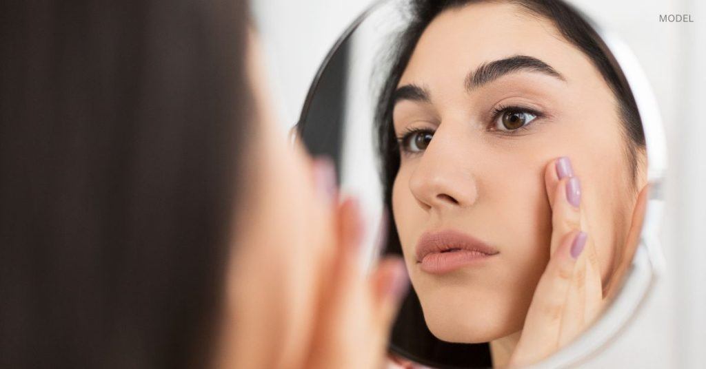 Woman with clear and radiant skin (model) examines her face in the mirror.
