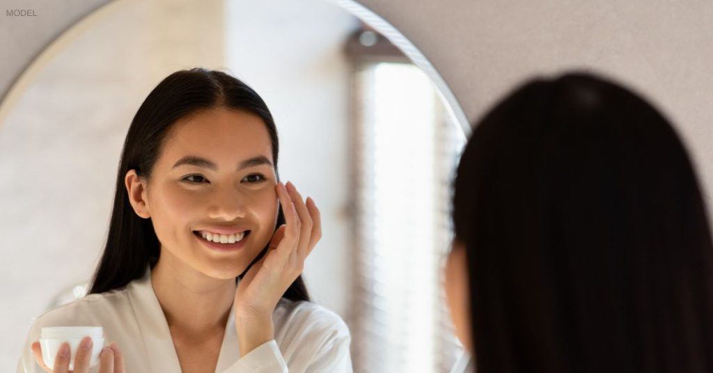 woman standing in mirror applying product on face (MODEL)